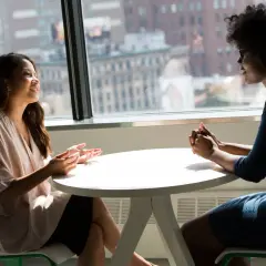 Dos mujeres conversan animadamente en una mesa. Una de ellas gesticula con las manos, mientras la otra escucha atentamente. Son **fotos speaking B2** de una conversación fluida.