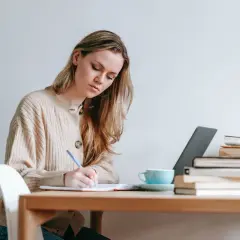 Mujer rubia concentrada escribiendo en un cuaderno, con libros y taza de café. Ideal para texto en inglés B1.