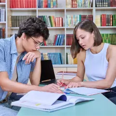 Dos personas jóvenes estudian juntas en una biblioteca, rodeadas de estanterías llenas de libros.