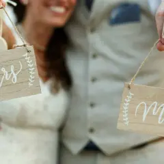 Novios sonrientes sostienen letreros de madera: "Mrs" y "Mr". ¡La señora es ahora parte de la pareja!