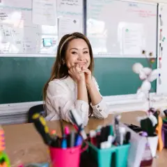 Sonríe una maestra en un aula, con pizarras detrás y materiales escolares en primer plano. Ella parece mirar hacia adelante, con un significado de esperanza.