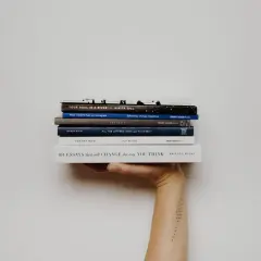 Mano sosteniendo una pila de libros, incluyendo "Your Soul Is a River" y "101 Essays that Will Change the Way You Think". Una lectura fascinante para cualquier aficionado a la novela inglesa.