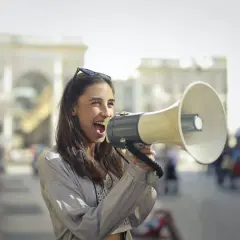 Mujer con megáfono, gritando con entusiasmo. Parece que está anunciando algo importante, como si dijera "¡Abran paso!" en inglés, usando un modismo para hacerse oír.
