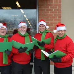 Cuatro hombres con gorros de Papá Noel y bufandas verdes cantan villancicos en inglés famosos, sosteniendo partituras verdes.