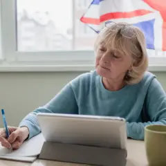 Mujer mayor estudia, con una tableta y taza, y la bandera del Reino Unido al fondo. Aprende inglés, o algo más.