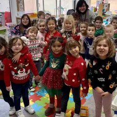 Niños y adultos celebran el jumper day con suéteres festivos en un aula. ¡Felices fiestas!
