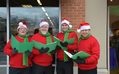 Cuatro hombres con gorros de Papá Noel y bufandas verdes cantan villancicos en inglés famosos, sosteniendo partituras verdes.