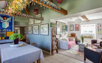 Interior de una casa típica inglesa con decoración colorida y vintage. Se ve un comedor con mantel a rayas y un salón con sofá, alfombra y cuadros.