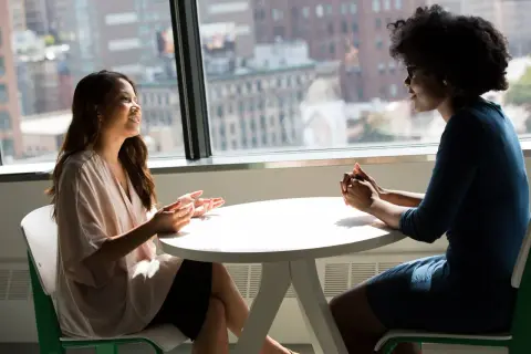 Dos mujeres conversan animadamente en una mesa. Una de ellas gesticula con las manos, mientras la otra escucha atentamente. Son **fotos speaking B2** de una conversación fluida.