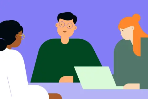 Tres people están reunidos, uno con gafas azules mira a la cámara, otro con cabello naranja mira una laptop.