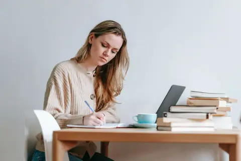 Mujer rubia concentrada escribiendo en un cuaderno, con libros y taza de café. Ideal para texto en inglés B1.