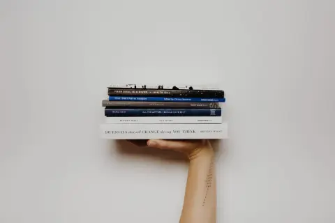 Mano sosteniendo una pila de libros, incluyendo "Your Soul Is a River" y "101 Essays that Will Change the Way You Think". Una lectura fascinante para cualquier aficionado a la novela inglesa.