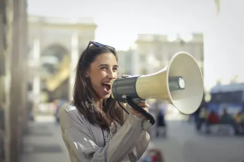 Mujer con megáfono, gritando con entusiasmo. Parece que está anunciando algo importante, como si dijera "¡Abran paso!" en inglés, usando un modismo para hacerse oír.