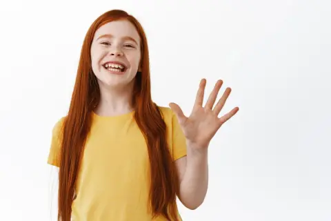 Niña pelirroja sonriente saluda con la mano, ¡how are ya!