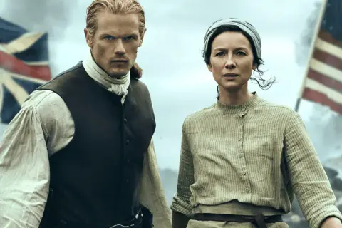 Jamie y Claire, protagonistas de series inglesas antiguas, se preparan para la batalla. Banderas ondean al fondo.