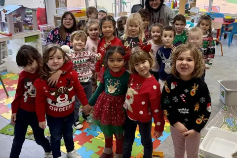Niños y adultos celebran el jumper day con suéteres festivos en un aula. ¡Felices fiestas!