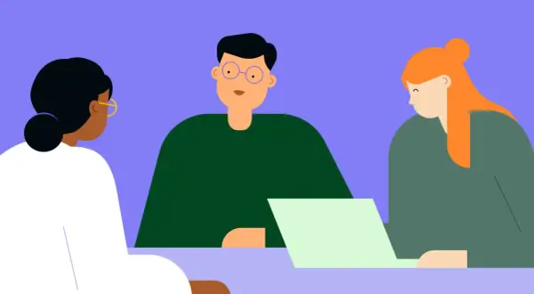 Tres people están reunidos, uno con gafas azules mira a la cámara, otro con cabello naranja mira una laptop.