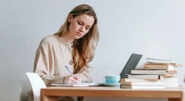 Mujer rubia concentrada escribiendo en un cuaderno, con libros y taza de café. Ideal para texto en inglés B1.