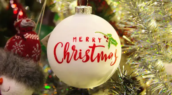 Adorno blanco con "Merry Christmas" en rojo. Un muñeco de nieve tejido y guirnaldas brillantes completan la escena festiva.