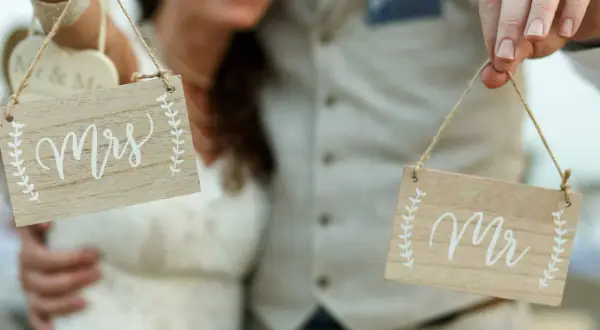 Novios sonrientes sostienen letreros de madera: "Mrs" y "Mr". ¡La señora es ahora parte de la pareja!