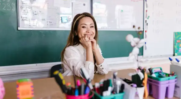 Sonríe una maestra en un aula, con pizarras detrás y materiales escolares en primer plano. Ella parece mirar hacia adelante, con un significado de esperanza.