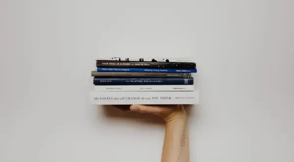 Mano sosteniendo una pila de libros, incluyendo "Your Soul Is a River" y "101 Essays that Will Change the Way You Think". Una lectura fascinante para cualquier aficionado a la novela inglesa.