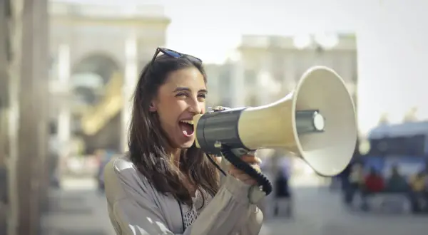 Mujer con megáfono, gritando con entusiasmo. Parece que está anunciando algo importante, como si dijera "¡Abran paso!" en inglés, usando un modismo para hacerse oír.