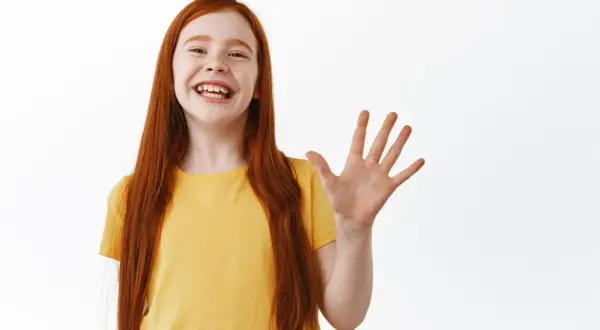 Niña pelirroja sonriente saluda con la mano, ¡how are ya!