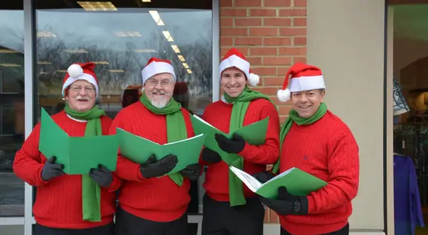 Cuatro hombres con gorros de Papá Noel y bufandas verdes cantan villancicos en inglés famosos, sosteniendo partituras verdes.