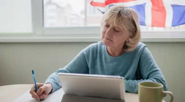Mujer mayor estudia, con una tableta y taza, y la bandera del Reino Unido al fondo. Aprende inglés, o algo más.