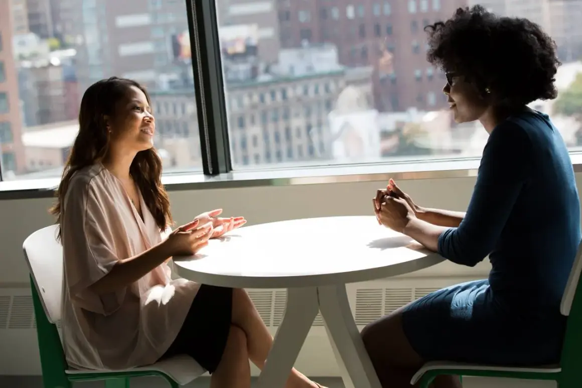 Dos mujeres conversan animadamente en una mesa. Una de ellas gesticula con las manos, mientras la otra escucha atentamente. Son **fotos speaking B2** de una conversación fluida.