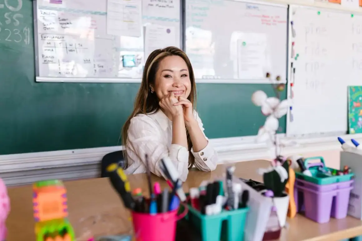 Sonríe una maestra en un aula, con pizarras detrás y materiales escolares en primer plano. Ella parece mirar hacia adelante, con un significado de esperanza.