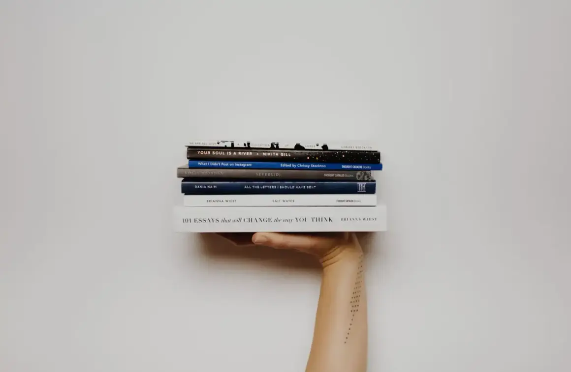 Mano sosteniendo una pila de libros, incluyendo "Your Soul Is a River" y "101 Essays that Will Change the Way You Think". Una lectura fascinante para cualquier aficionado a la novela inglesa.