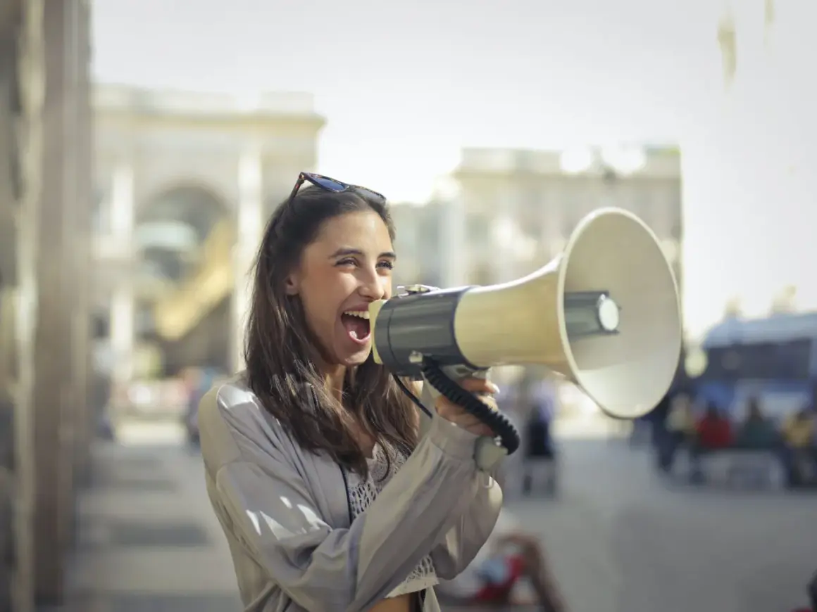 Mujer con megáfono, gritando con entusiasmo. Parece que está anunciando algo importante, como si dijera "¡Abran paso!" en inglés, usando un modismo para hacerse oír.
