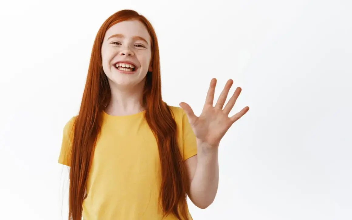 Niña pelirroja sonriente saluda con la mano, ¡how are ya!