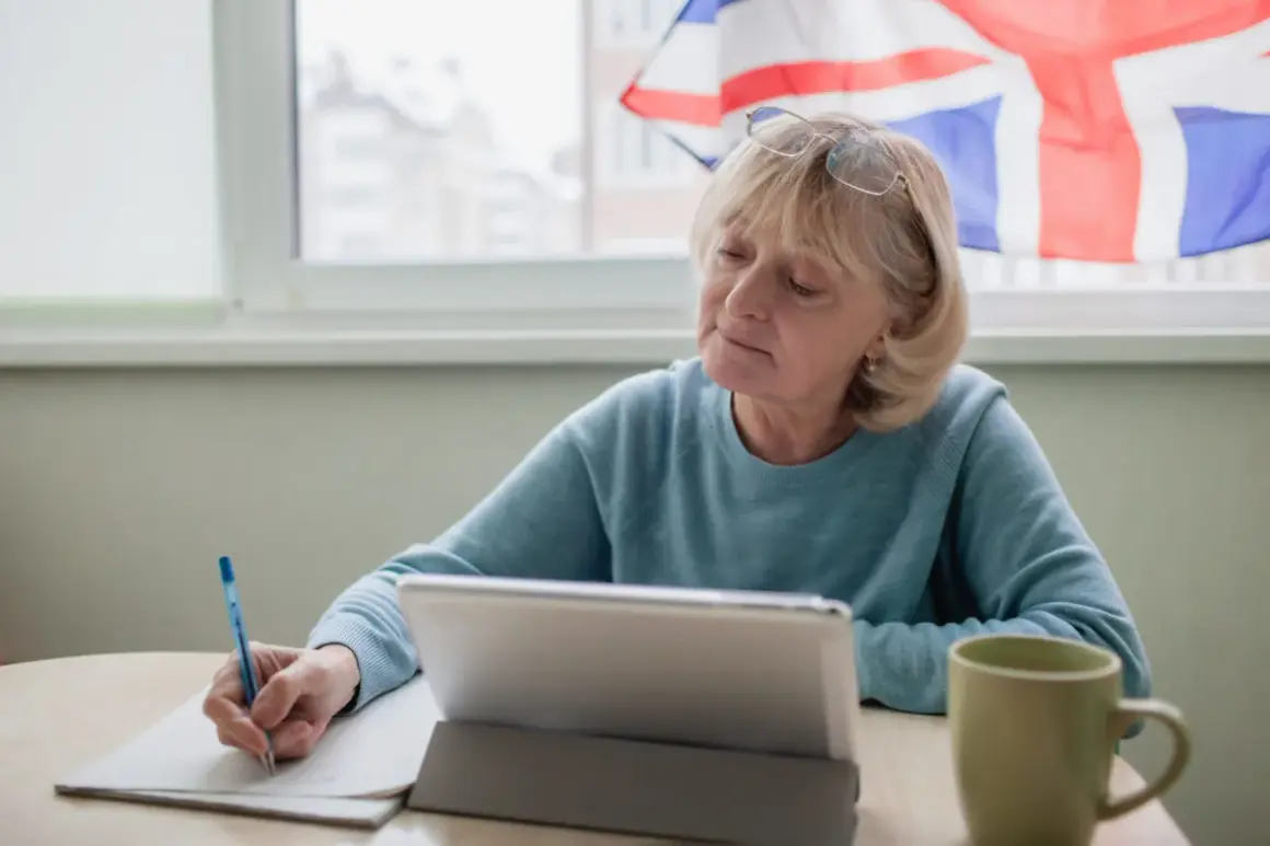 Mujer mayor estudia, con una tableta y taza, y la bandera del Reino Unido al fondo. Aprende inglés, o algo más.