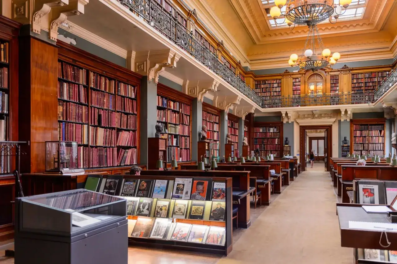 Interior de una biblioteca majestuosa, con estanterías repletas de libros que evocan clásicos de la literatura. Lámparas de lectura y bustos adornan los pupitres.
