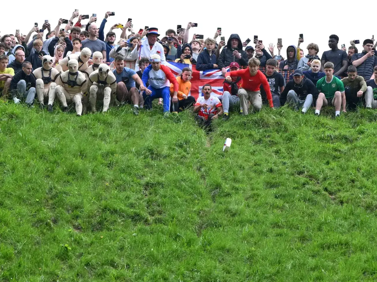 Participantes disfrazados y espectadores esperan el inicio del cheese rolling en una colina verde.