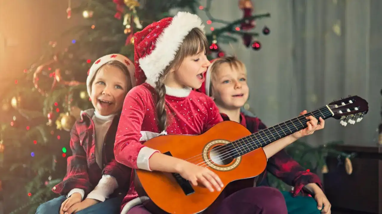 Niños cantan un villancico en inglés junto a un árbol de Navidad, uno toca la guitarra.