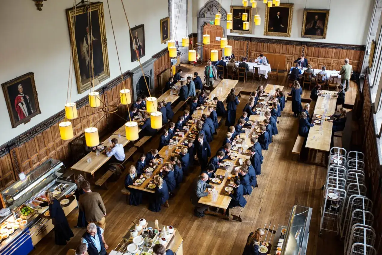 Comedor de internados en Reino Unido. Estudiantes comen en largas mesas de madera bajo lámparas amarillas, rodeados de retratos históricos.