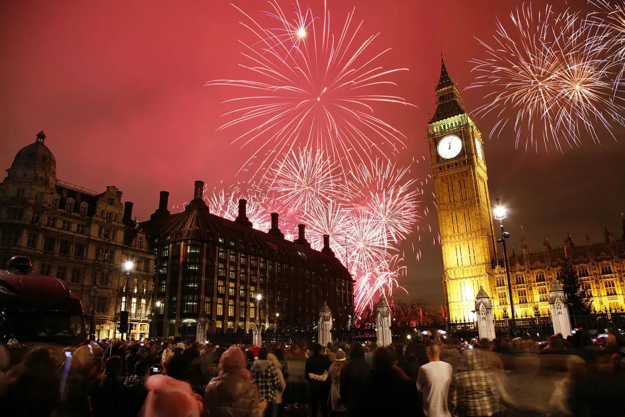 Fuegos artificiales iluminan el cielo sobre el Big Ben y el Parlamento, celebrando las tradiciones de Londres. Una multitud observa el espectáculo.