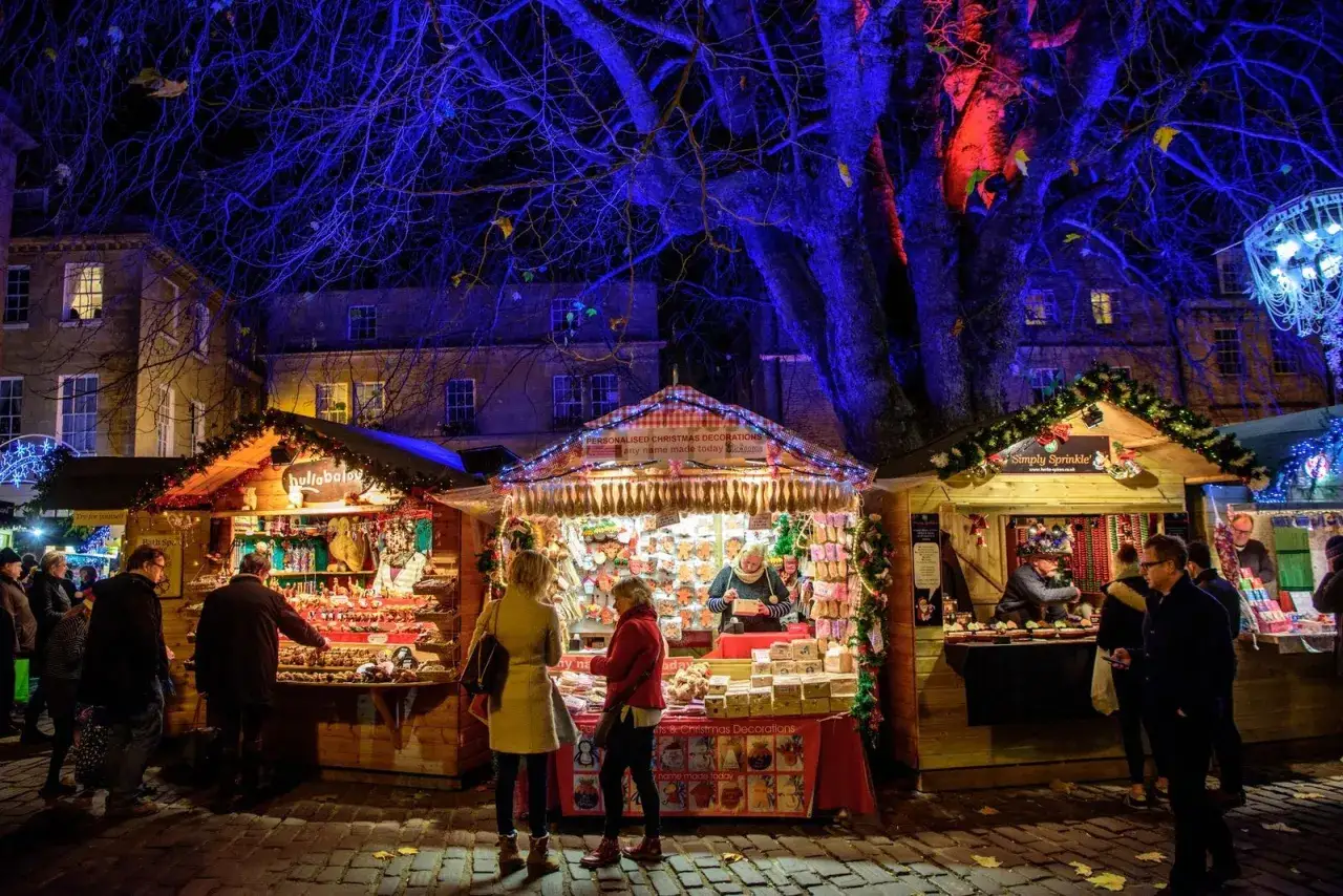 Mercado navideño en Gran Bretaña, con puestos iluminados y gente comprando decoraciones. El árbol azul ilumina la escena.