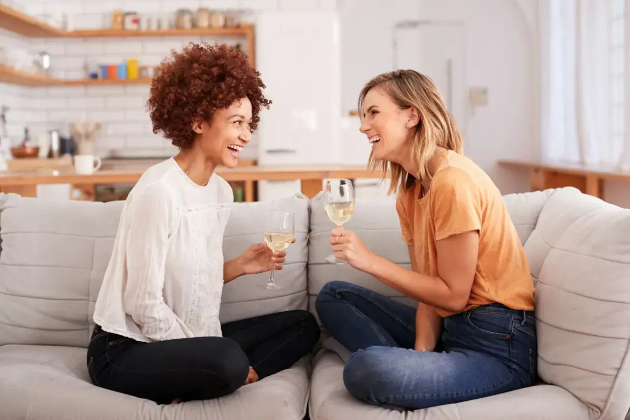 Dos amigas brindan con vino blanco en un sofá, riendo y preguntándose