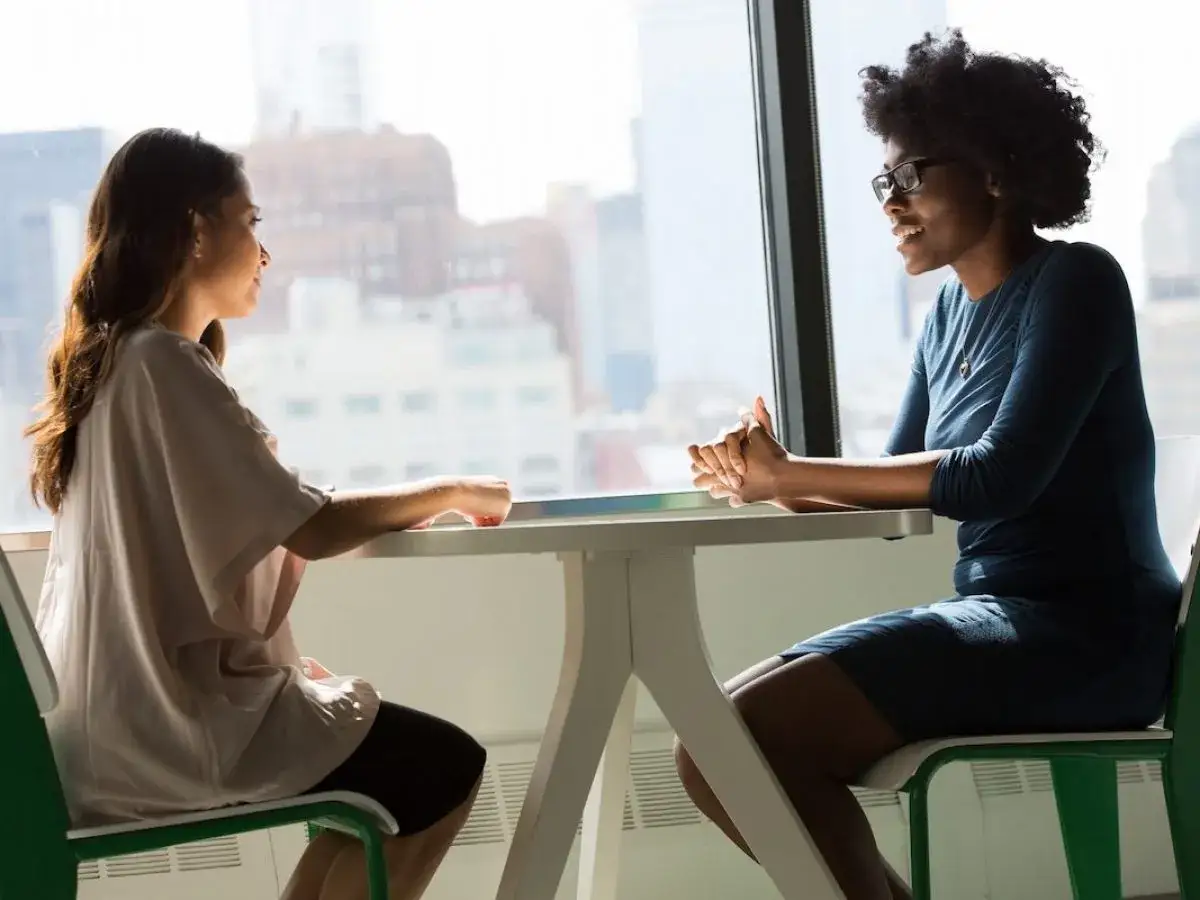 Dos mujeres conversan animadamente. Una, con gafas, parece contar una anécdota divertida, como si su mascota estuviera hablando.