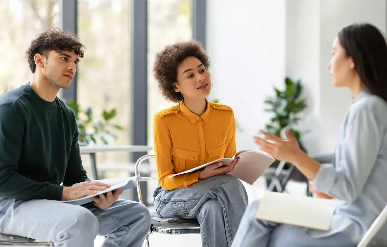 Tres personas en un círculo, discutiendo y tomando notas. Una mujer con pelo afro y camisa naranja sostiene un libro abierto. Son fotos speaking B2.