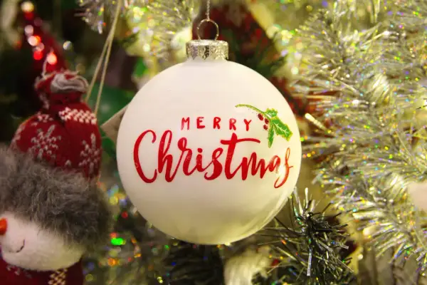 Adorno blanco con "Merry Christmas" en rojo. Un muñeco de nieve tejido y guirnaldas brillantes completan la escena festiva.