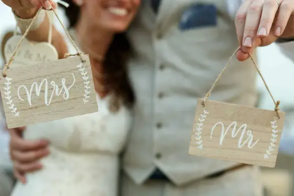 Novios sonrientes sostienen letreros de madera: "Mrs" y "Mr". ¡La señora es ahora parte de la pareja!